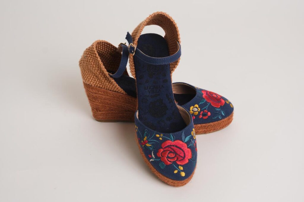 Encantadoras alpargatas de cuña en azul marino, decoradas con un colorido bordado floral que destaca rosas rojas en la puntera. Diseñadas con pulsera ajustable y acabado en esparto, ofrecen un equilibrio perfecto entre tradición flamenca y modernidad, siendo ideales para un estilo elegante y cómodo.