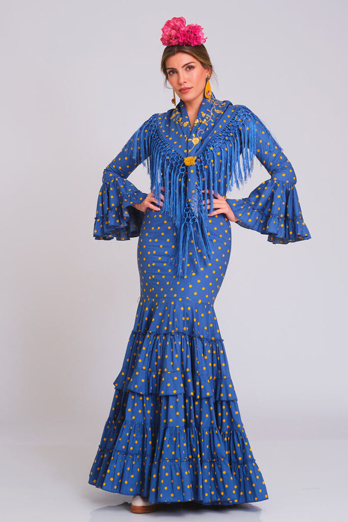 Vestido de flamenca en un intenso tono azul con alegre estampado de lunares amarillos. Diseño de corte sirena que entalla la figura, con una falda de volantes sucesivos que aporta gran movimiento y un estilo tradicional muy característico.