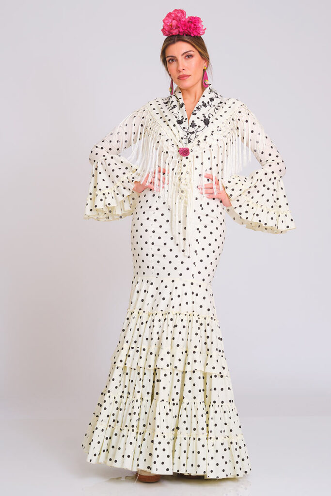 Elegante traje de flamenca en color blanco con clásico estampado de lunares negros. Diseño de corte sirena que entalla la figura con distinción, complementado con una falda de volantes sucesivos que aporta un movimiento dinámico y una estética tradicional muy estilizada.