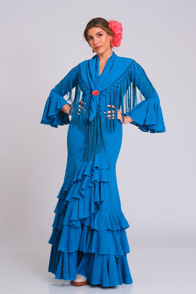 Diseño de flamenca en un vibrante tono azul con delicada textura de lunares. Corte sirena que define la silueta con elegancia, acompañado de una falda de volantes en cascada que aporta un movimiento fluido y auténtico para lucir con distinción.