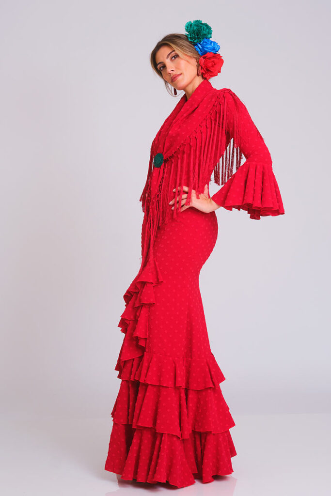 Traje de flamenca en color rojo intenso, caracterizado por una sutil textura de lunares. Diseño de corte sirena que entalla la figura con elegancia, culminando en una falda de volantes sucesivos que brinda un movimiento dinámico y una estética clásica para eventos de moda flamenca.