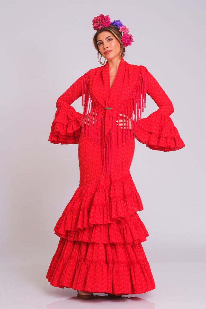 Apasionado vestido de flamenca en color rojo intenso con sutil textura de lunares. Diseño de corte sirena que entalla la figura, complementado con una falda de volantes en cascada para un movimiento dinámico y una estética clásica renovada.