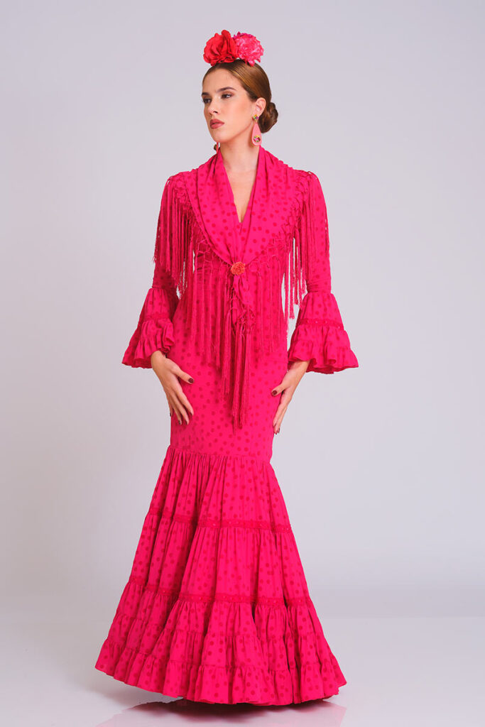 Traje de flamenca en un vibrante tono fucsia con sutil estampado de lunares. Corte sirena que entalla la silueta con elegancia, presentando una falda de volantes sucesivos que proporciona un movimiento dinámico y una estética renovada para tus eventos especiales.