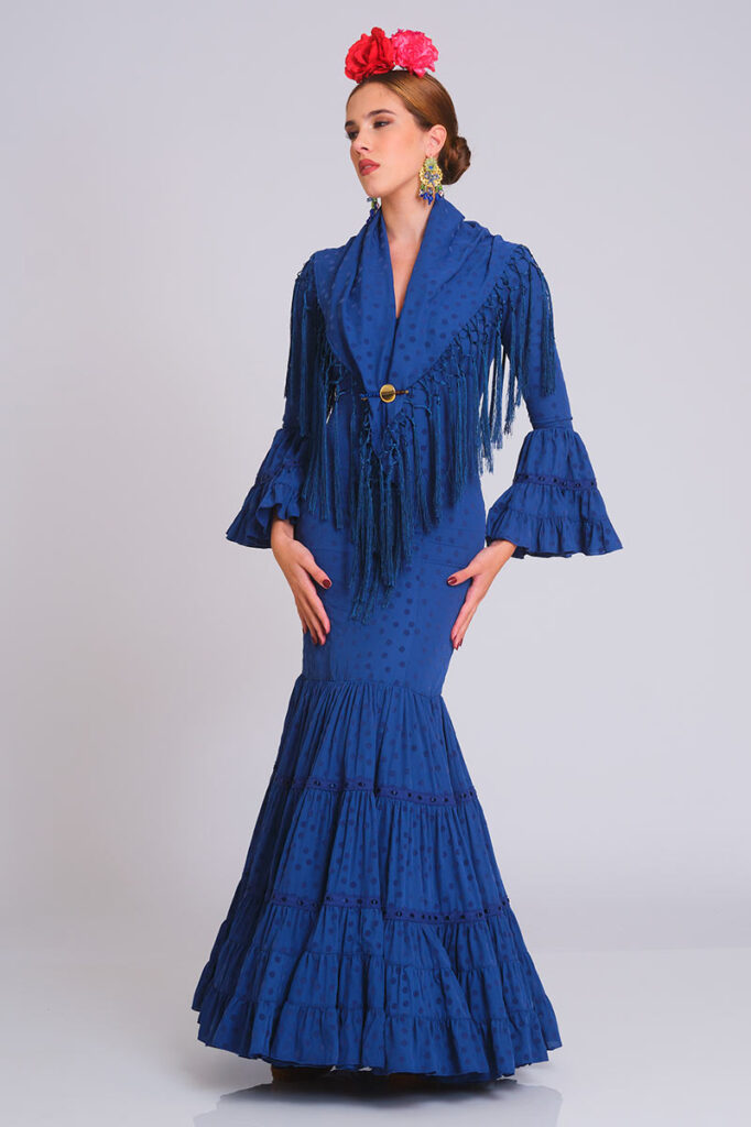 Diseño de flamenca en un profundo azul marino con estampado de lunares tono sobre tono. Corte sirena que se ajusta perfectamente a la figura, con falda de volantes sucesivos ribeteados para un acabado clásico y elegante.