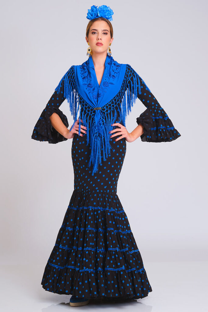 Este vestido de flamenca destaca por su elegante combinación de fondo negro con lunares en azul eléctrico. Su corte sirena entalla la silueta, mientras que la falda de volantes sucesivos, acentuada con ribetes en azul, aporta un movimiento dinámico y un contraste visual sofisticado y muy actual.