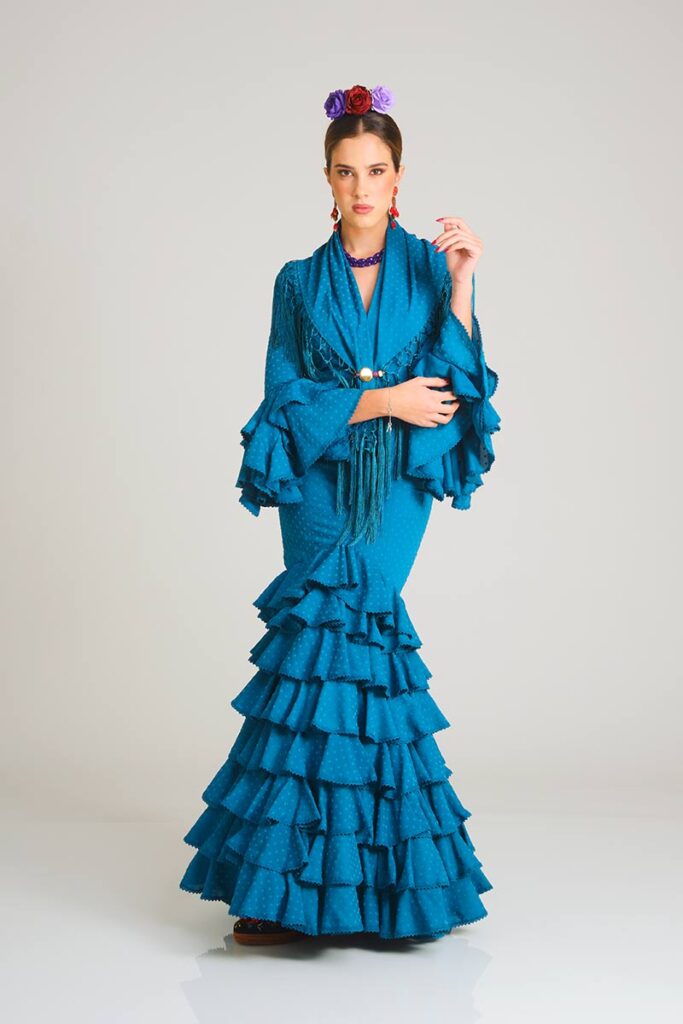 Este sofisticado vestido de flamenca en un profundo tono azul cerceta presenta un sutil estampado de lunares tono sobre tono. Su diseño de corte sirena entalla la silueta con elegancia, mientras que la falda, estructurada en una cascada de volantes, proporciona un movimiento fluido y una estética tradicional renovada para tus eventos especiales.