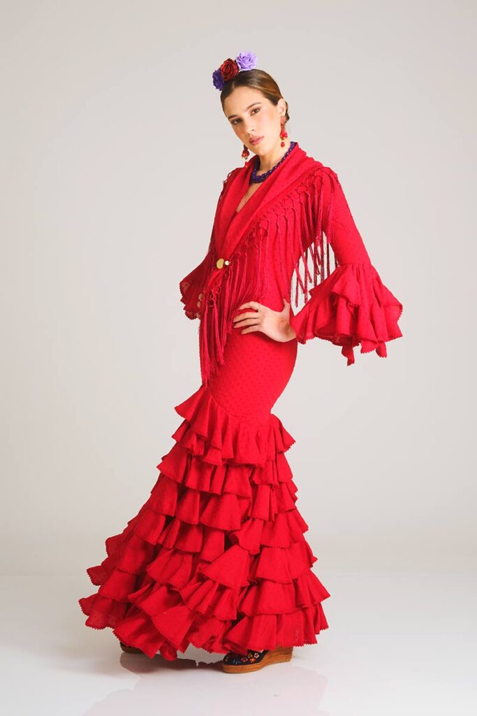 Este apasionado vestido de flamenca en un rojo intenso destaca por su sutil textura de lunares. El diseño entallado de corte sirena realza la silueta, mientras que su falda de volantes sucesivos aporta una caída espectacular y un movimiento lleno de fuerza para tus ocasiones especiales.