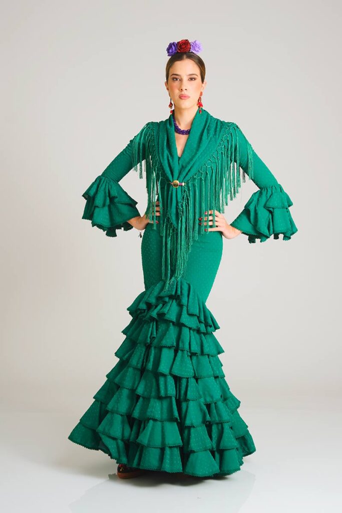 Este vestido de flamenca en un intenso tono verde esmeralda presenta un sutil estampado de lunares tono sobre tono. Su diseño de corte sirena entalla la silueta con elegancia, mientras que la falda, estructurada en una cascada de volantes, aporta un movimiento dinámico y una estética tradicional renovada, ideal para destacar en cualquier ocasión.