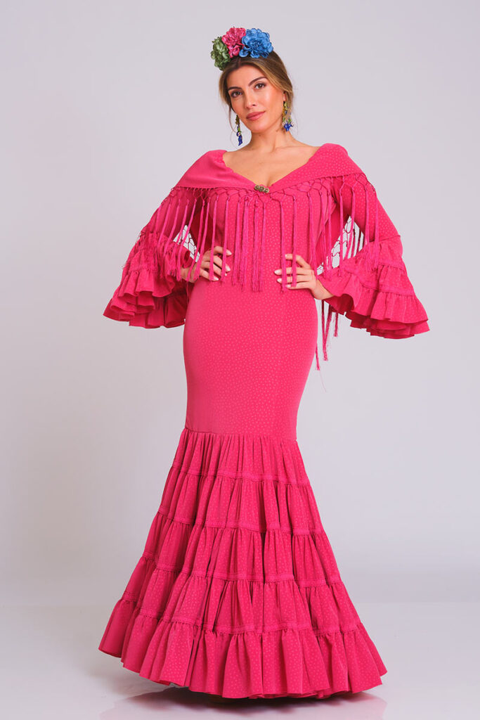 Este vibrante vestido de flamenca en color fucsia presenta un delicado estampado de minilunares al tono. Destaca por su favorecedor corte sirena que estiliza la figura y una falda compuesta por sucesivos volantes con detalles de encaje, ideal para lucir con elegancia en cualquier evento.