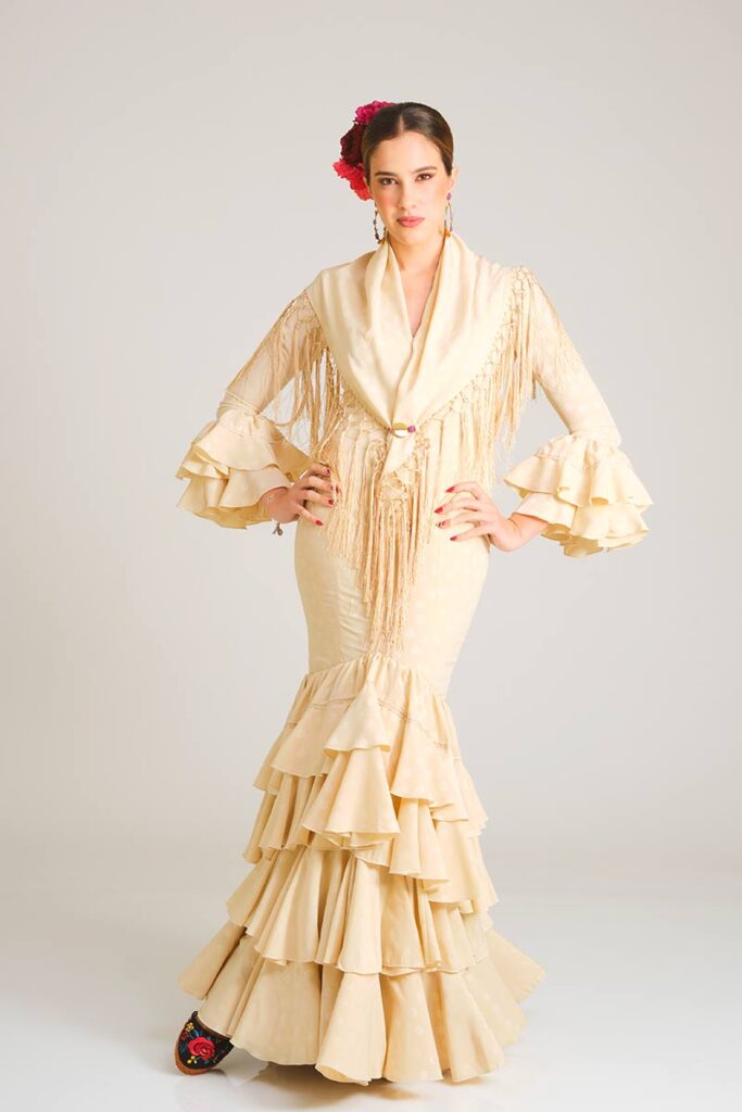 Este elegante vestido de flamenca en un suave tono crema presenta un delicado estampado de lunares tono sobre tono, aportando un aire refinado y minimalista. Su diseño de corte sirena realza la silueta con sutileza, mientras que la falda, estructurada en una cascada de volantes, proporciona un movimiento fluido y auténtico que captura la esencia de la tradición flamenca.