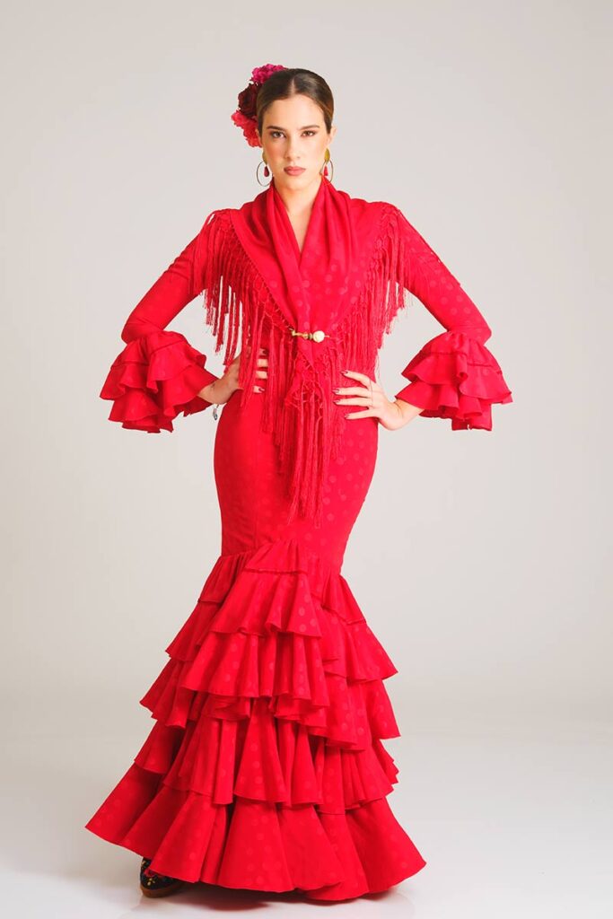 Este vestido de flamenca en un vibrante tono rojo presenta un elegante estampado de lunares al mismo tono. Su diseño entallado de corte sirena realza la silueta, mientras que la falda, estructurada en múltiples volantes, aporta un movimiento espectacular y una estética profundamente tradicional.