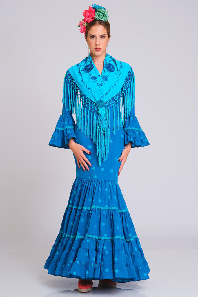 Este cautivador vestido de flamenca en un intenso tono azul turquesa luce un estampado de lunares en un azul ligeramente más claro. Su diseño entallado de corte sirena realza la silueta con gracia, mientras que la falda, formada por capas de volantes decoradas con encaje, aporta una sensación de movimiento dinámico y tradicional.