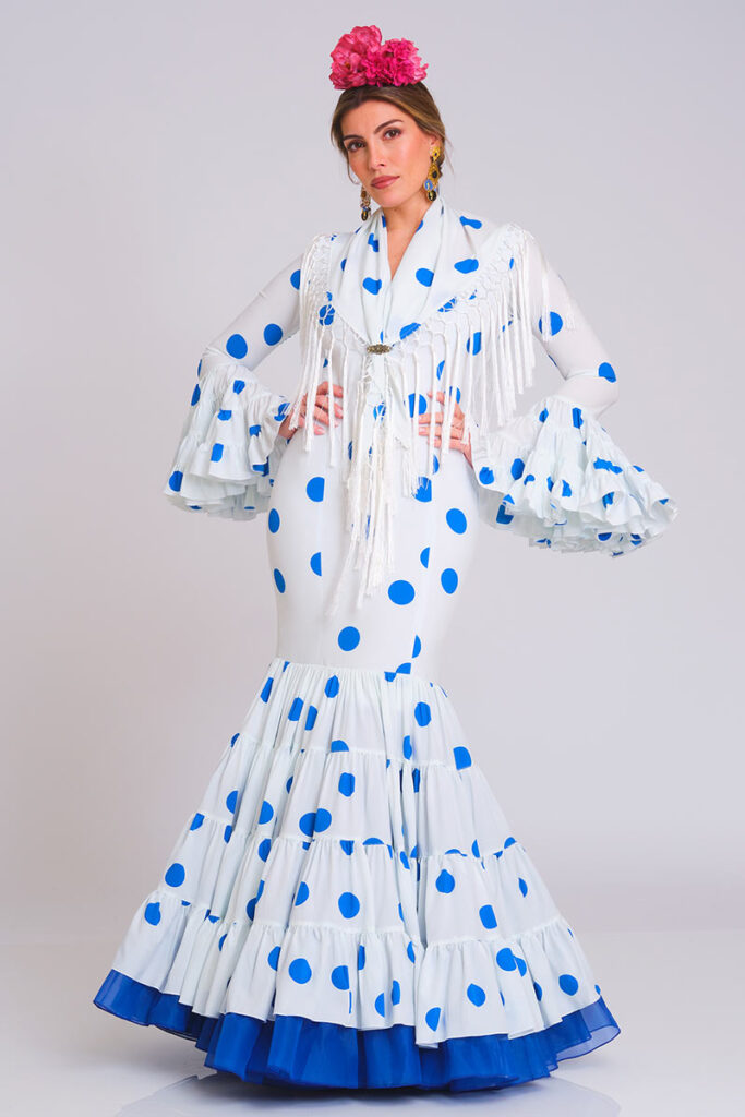 Este vestido de flamenca destaca por su combinación de blanco puro con lunares azul vibrante, ofreciendo una estética fresca y llamativa. El diseño presenta un corte sirena que estiliza la figura y una falda estructurada en volantes, rematada con un borde inferior en contraste que añade elegancia al conjunto.