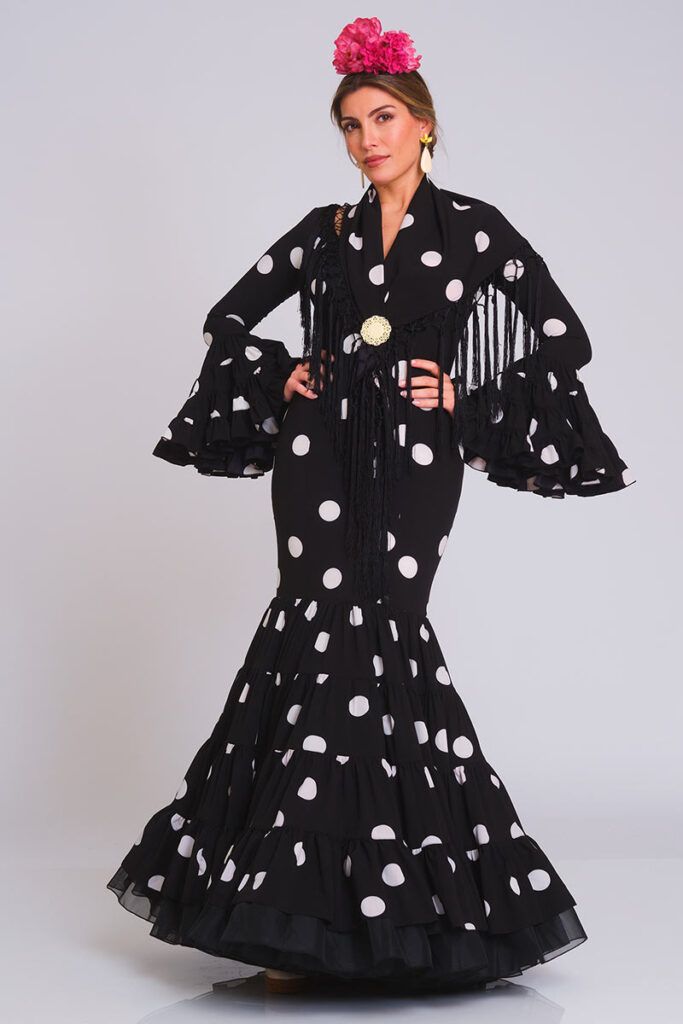 Este elegante vestido de flamenca combina un clásico fondo negro con lunares blancos, realzado por un corte sirena que aporta sofisticación y una falda estructurada con volantes que garantiza un movimiento fluido al caminar.