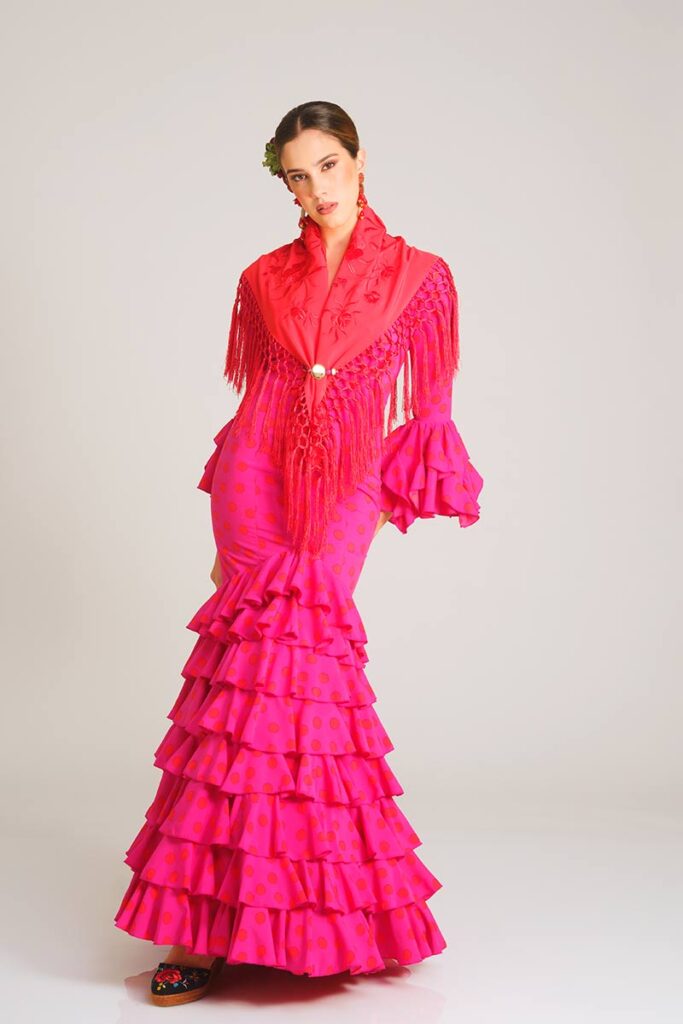Vestido de flamenca en un intenso tono fucsia con lunares rojos, diseñado con un corte sirena que ajusta la figura y una falda estructurada en volantes sucesivos, creando una imagen llena de fuerza y estilo tradicional.