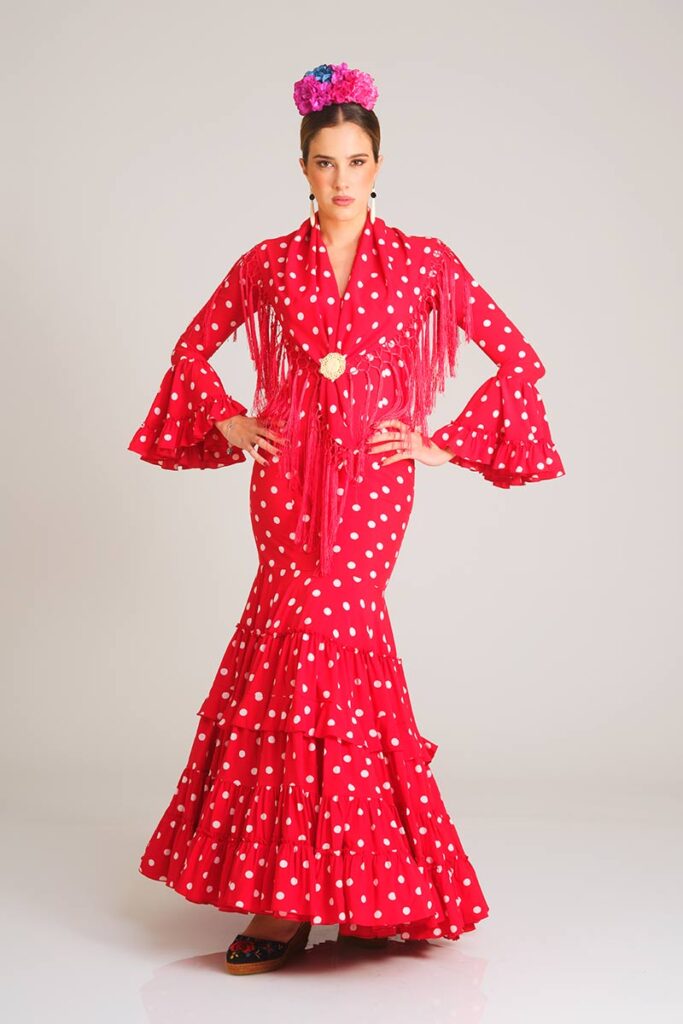 Vestido de flamenca con un alegre estampado de lunares blancos sobre fondo rojo, diseñado con un corte sirena entallado que culmina en una falda de amplios volantes, ideal para lucir un estilo folclórico y muy femenino.