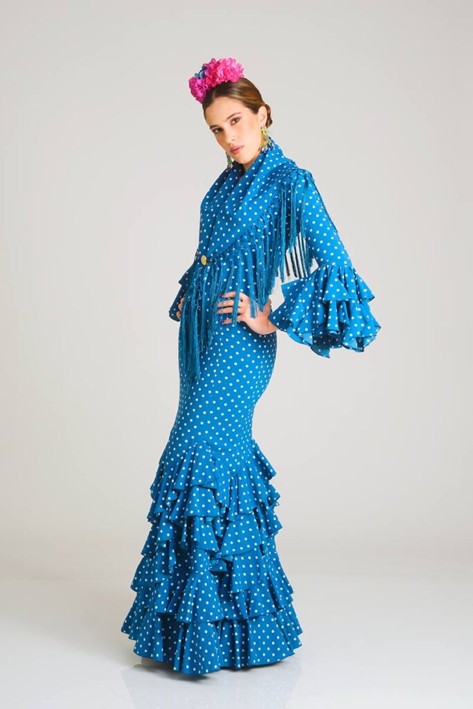 Traje de flamenca diseñado en un tono azul vibrante con estampado de lunares blancos, destacando por un corte sirena que ajusta la figura y una falda ornamentada con volantes superpuestos para lograr una apariencia clásica y refinada.