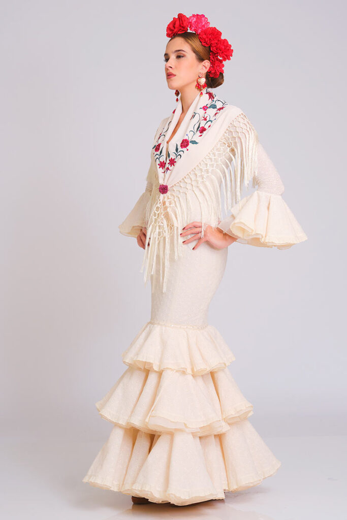 Vestido de flamenca en color crema suave con sutil estampado de lunares, diseñado con cuerpo ajustado y falda larga de volantes escalonados. Traje de gitana artesanal que destaca por su mantoncillo bordado en tonos florales con flecos largos y mangas de doble volante. Pieza elegante de alta costura, ideal para lucir en la Feria de Abril, romerías y eventos de moda andaluza