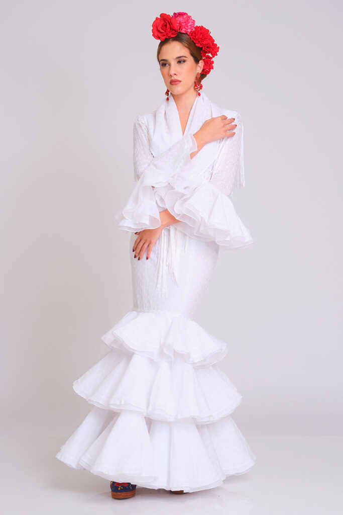 Vestido de flamenca en color blanco puro con sutil estampado de lunares, diseñado con cuerpo ajustado y falda larga de volantes escalonados. Traje de gitana artesanal que destaca por su mantoncillo blanco a juego con flecos largos y mangas de doble volante. Pieza elegante de alta costura, ideal para lucir en la Feria de Abril, romerías y eventos de moda andaluza