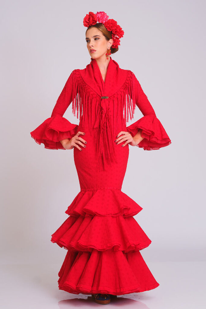 Vestido de flamenca en color rojo intenso con sutil estampado de lunares, diseñado con cuerpo ajustado y falda larga de volantes escalonados. Traje de gitana artesanal que destaca por su mantón a juego con flecos largos y mangas de doble volante. Pieza elegante de alta costura, ideal para lucir en la Feria de Abril, romerías y eventos de moda andaluza