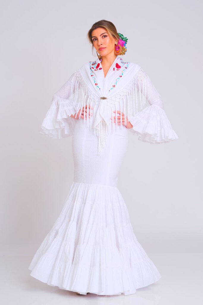 Vestido de flamenca en color blanco impoluto con sutil estampado de lunares, diseñado con cuerpo ajustado y falda larga de volantes escalonados. Traje de gitana artesanal que destaca por su mantoncillo bordado en tonos florales con flecos largos y mangas de doble volante. Pieza elegante de alta costura, ideal para lucir en la Feria de Abril, romerías y eventos de moda andaluza