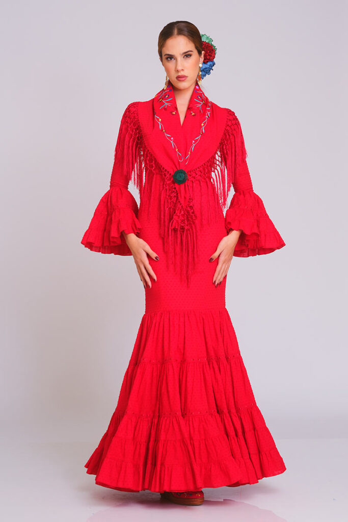 Vestido de flamenca rojo vibrante con cuerpo ajustado, falda de volantes en cascada y estampado de lunares sutil. Traje de gitana artesanal combinado con mantón bordado a juego y flecos largos. Mangas de doble volante. Diseño elegante de alta costura, perfecto para la Feria de Abril, romerías y eventos de moda andaluza