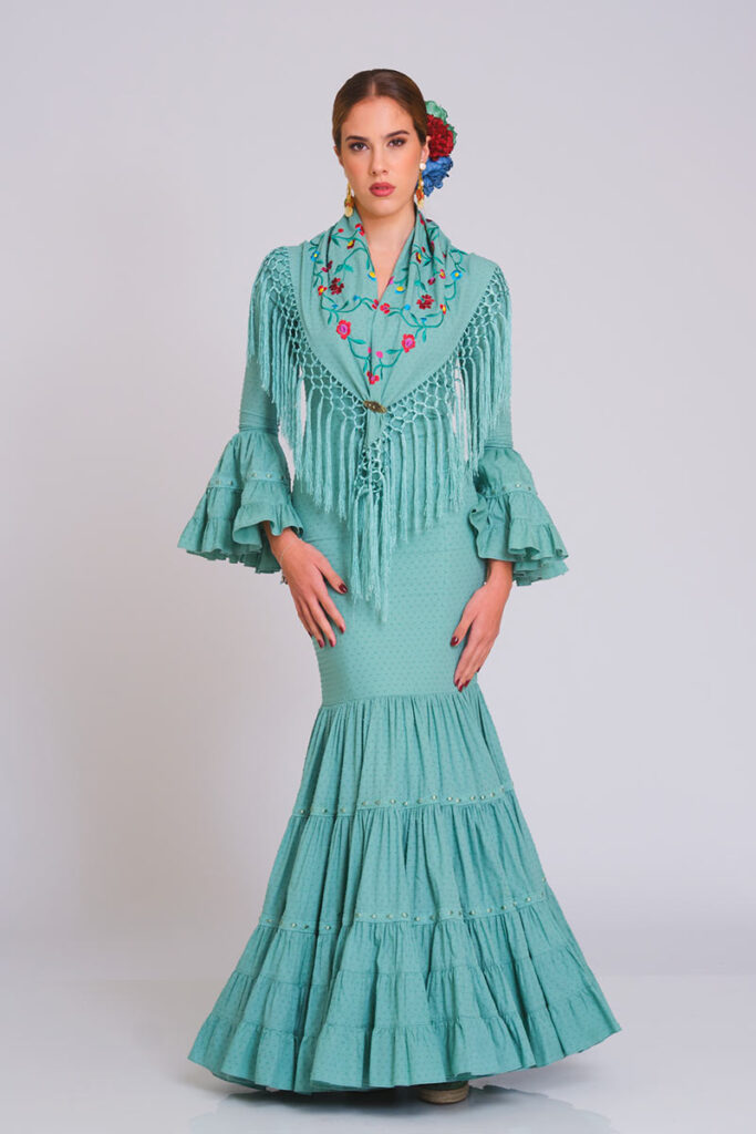 Vestido de flamenca en tono verde menta con estampado de lunares en relieve, corte sirena y falda de volantes escalonados. Traje de gitana artesanal para mujer, combinado con un mantoncillo bordado en motivos florales con flecos largos y mangas de doble volante. Diseño elegante de alta costura, ideal para la Feria de Abril, romerías y eventos flamencos