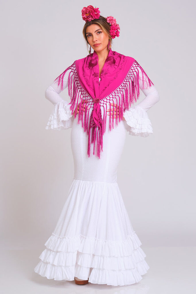 Vestido de flamenca blanco con estampado de lunares en relieve y corte sirena, presentando una falda de volantes escalonados. Traje de gitana para mujer, de estilo clásico y artesanal, combinado con un mantón de Manila en color fucsia vibrante con flecos largos. Mangas de doble volante. Perfecto para la Feria de Abril, romerías y eventos de moda flamenca