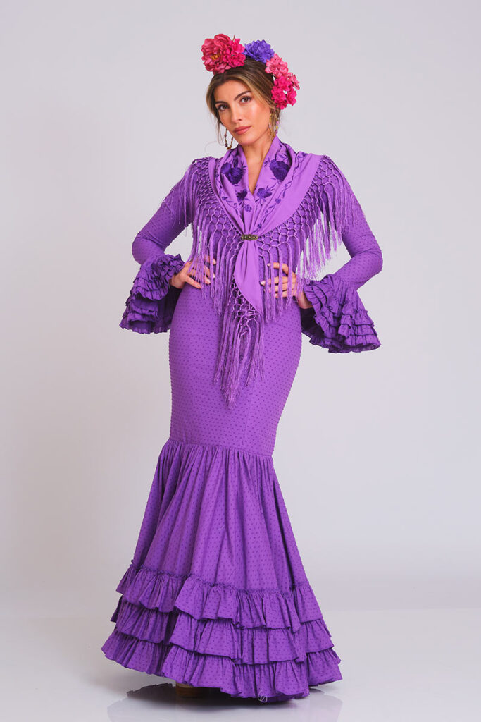 Vestido de flamenca morado con estampado sutil de lunares, cuerpo ajustado y falda de volantes en cascada. Traje de gitana artesanal para mujer, combinado con mantón bordado a juego con flecos largos. Mangas de doble volante. Diseño elegante ideal para la Feria de Abril, romerías y eventos de moda flamenca