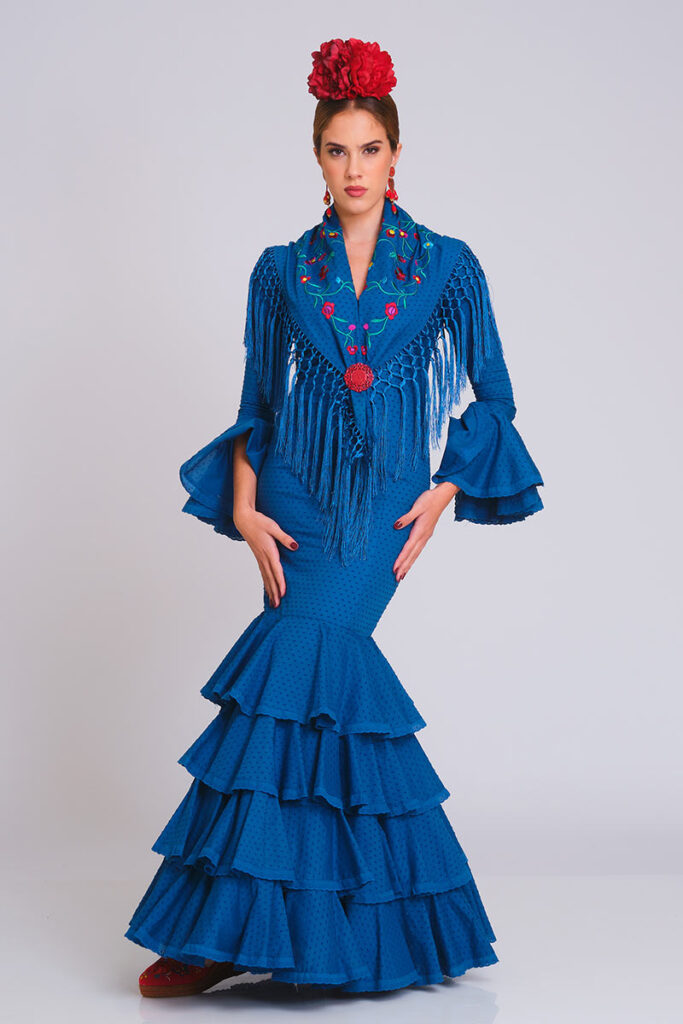 Alt Text: Vestido de flamenca azul intenso con estampado sutil de lunares y falda de volantes en cascada. Traje de gitana artesanal combinado con mantón bordado en tonos florales y flecos largos. Mangas con doble volante. Diseño elegante de alta costura, perfecto para lucir en la Feria de Abril, romerías y eventos de moda flamenca