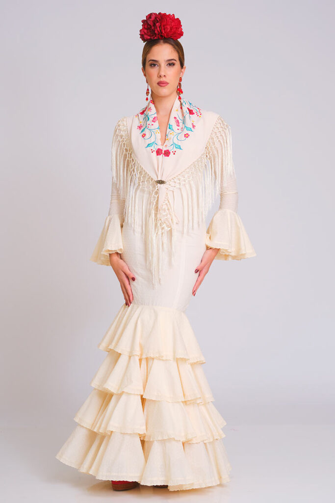 Vestido de flamenca color crema con falda de volantes en cascada y cuerpo ajustado. Traje de gitana para mujer, estilo clásico y elegante, combinado con mantón de Manila beige bordado con motivos florales y flecos largos. Mangas con doble volante. Perfecto para la Feria de Abril, romerías y eventos de moda flamenca
