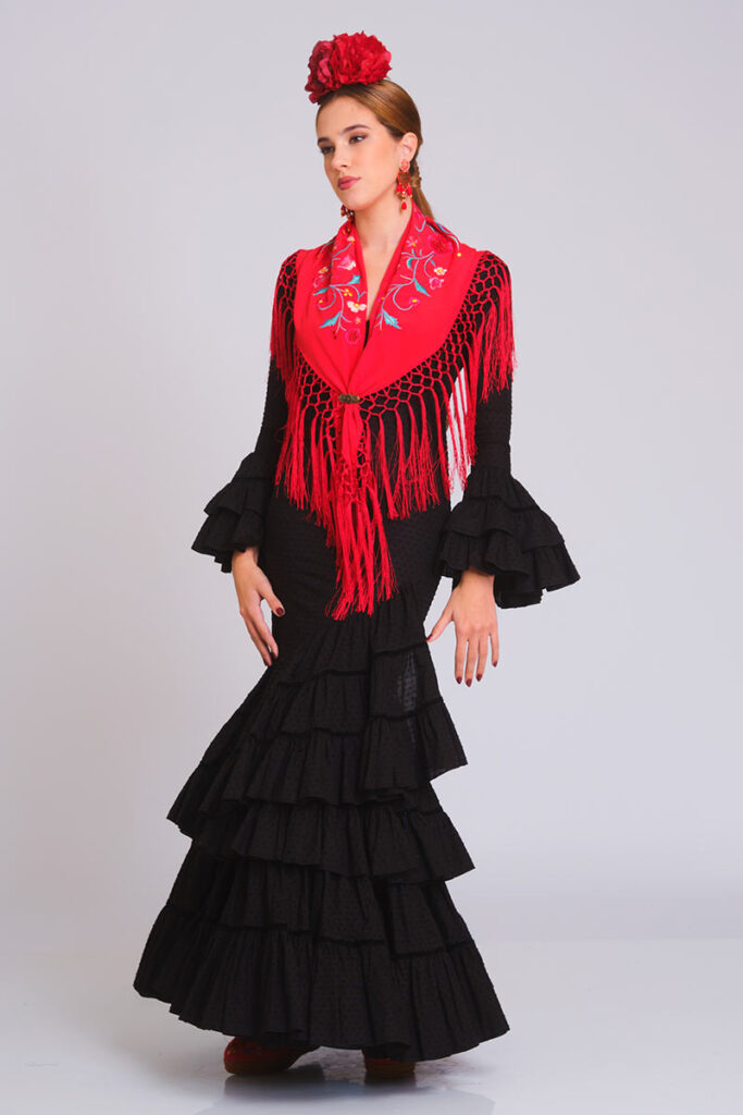 Vestido de flamenca negro con cuerpo ajustado y falda de volantes en capas, estilo clásico y elegante. Traje de gitana para mujer combinado con mantón de Manila rojo bordado con motivos florales y flecos largos. Mangas largas con volantes escalonados. Ideal para Feria de Abril, Romerías y espectáculos de danza española
