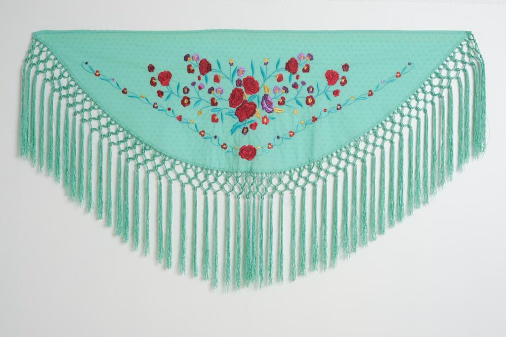 Exquisito mantón de flamenca triangular en color verde agua con textura de plumeti, ricamente bordado con un ramo central de rosas rojas y flores moradas. El diseño se completa con un festón de flecos largos y anudados en color a juego, creando un complemento tradicional y elegante para la moda andaluza