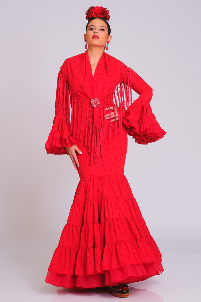 Clásico traje de flamenca en color rojo con textura de lunares, elaborado con un corte entallado que realza la figura y volantes fluidos en mangas y falda