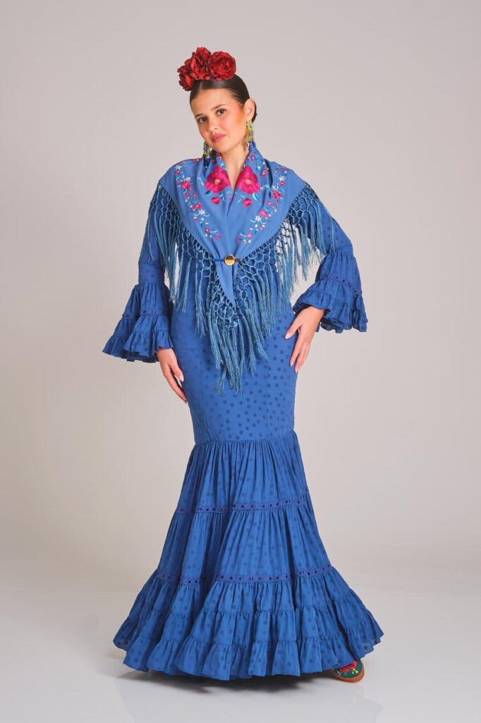 Vestido de flamenca en azul con estampado sutil de lunares, diseñado con un corte entallado que destaca por el volumen y movimiento de sus volantes, una elección elegante y tradicional para cualquier feria
