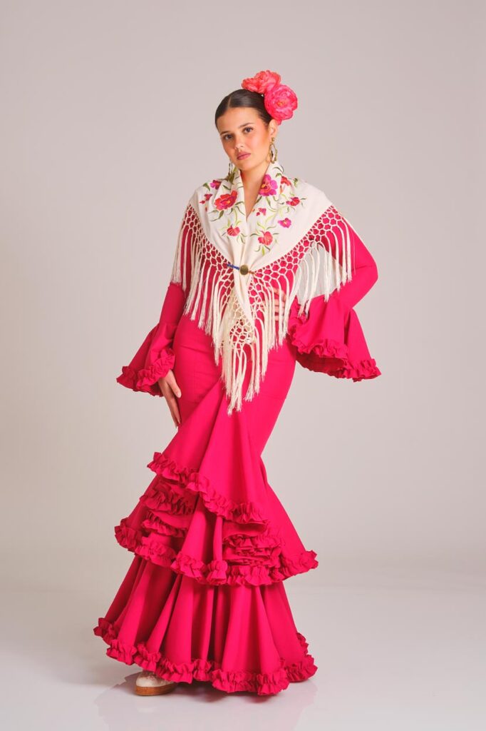 Traje de flamenca largo rojo elegante con falda de vuelos, combinado con un mantón blanco de bordado floral y flecos, un diseño clásico y vistoso para la feria.