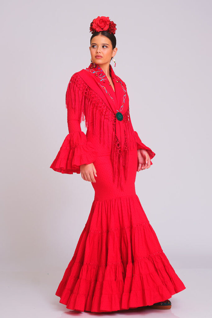 Vestido de flamenca rojo de corte sirena con mantoncillo bordado, flecos largos, mangas de volantes y flor roja en el cabello