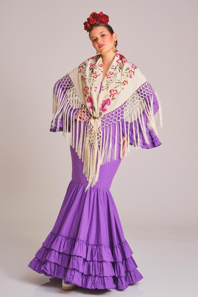 Modelo con vestido de flamenca morado de corte sirena, mantón marfil bordado, flecos largos y volantes en la falda
