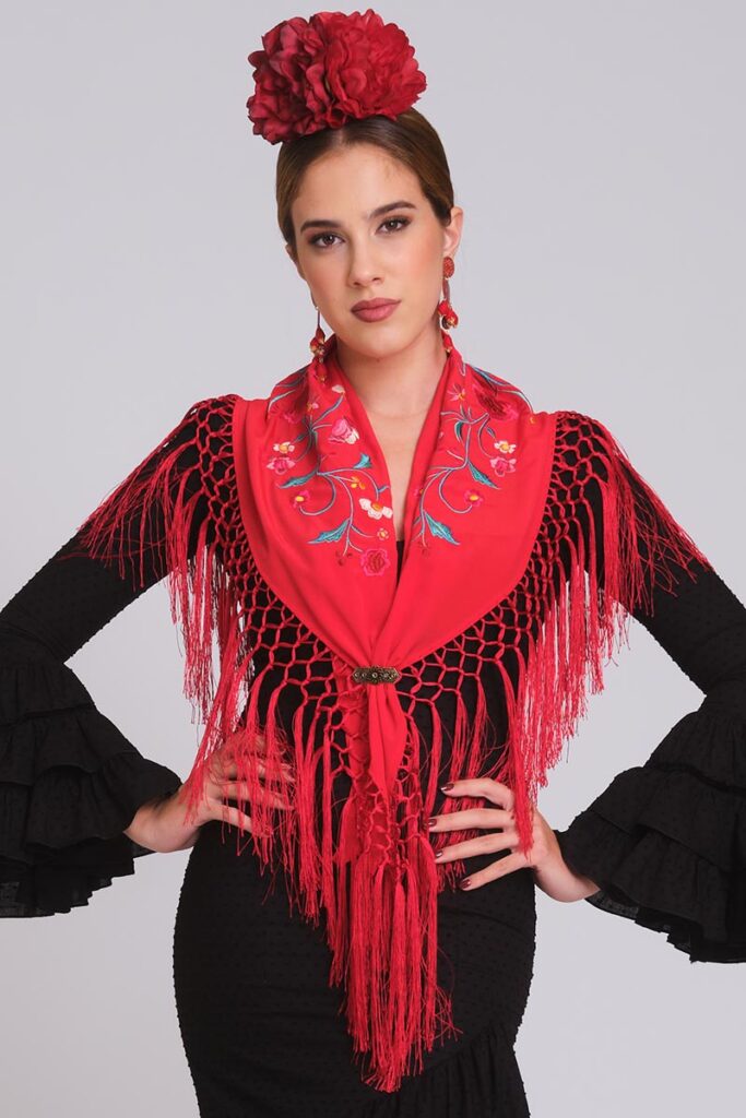 Modelo con traje de flamenca negro y mantoncillo rojo bordado, flecos artesanales, mangas de volantes y tocado floral