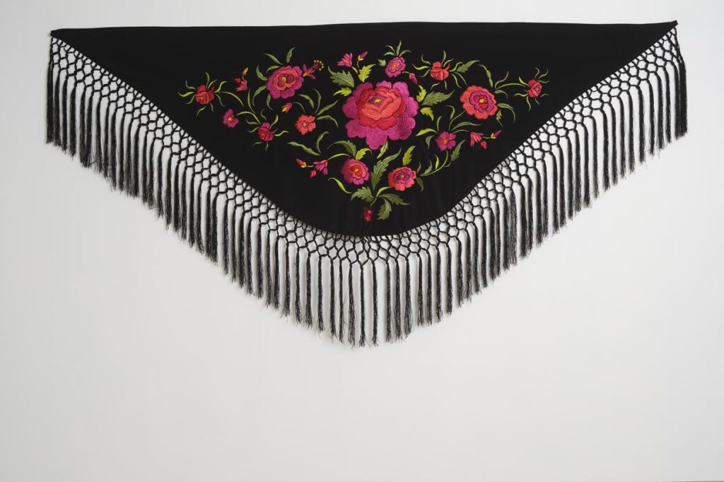 Mantón negro de moda flamenca con bordado floral fucsia y coral, hojas verdes y flecos largos anudados
