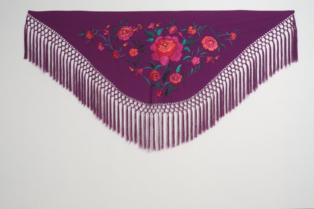 Mantón morado de moda flamenca con bordado floral en tonos coral y fucsia, hojas verdes y flecos largos de macramé