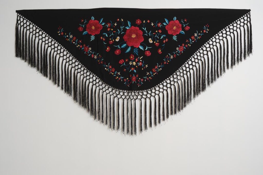 Mantón negro de moda flamenca con rosas rojas bordadas, hojas turquesa y flecos largos de macramé
