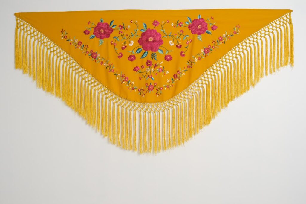 Mantón amarillo mostaza de moda flamenca con rosas fucsias bordadas, ramas multicolor y flecos largos anudados