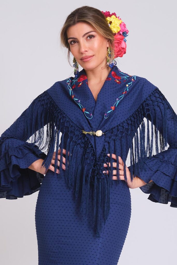 Moda flamenca en azul marino con mantoncillo bordado, flecos largos, mangas de volantes y flores en el cabello