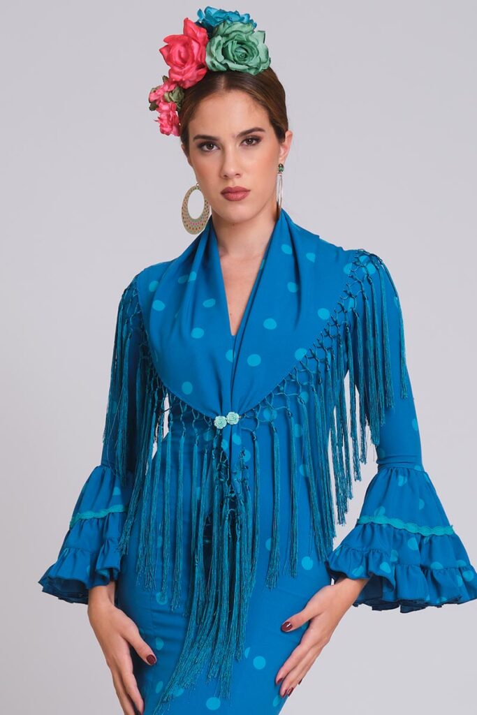 Vestido de flamenca azul pavo con lunares turquesa, mangas con volantes y mantoncillo de flecos largos cruzado.