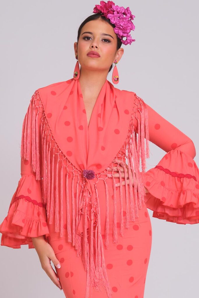 Vestido de flamenca coral con lunares rojos, mangas con volantes dobles y mantoncillo de flecos largos a juego.