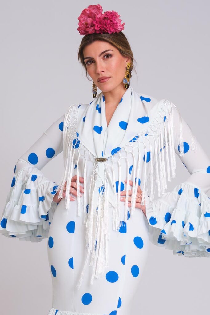 Traje de flamenca blanco con lunares azules, mangas de volantes y mantoncillo de flecos blanco cruzado con broche.