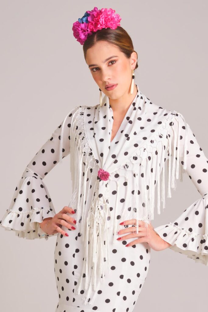Vestido de flamenca blanco con lunares negros, mantoncillo de flecos y flor rosa para look de feria