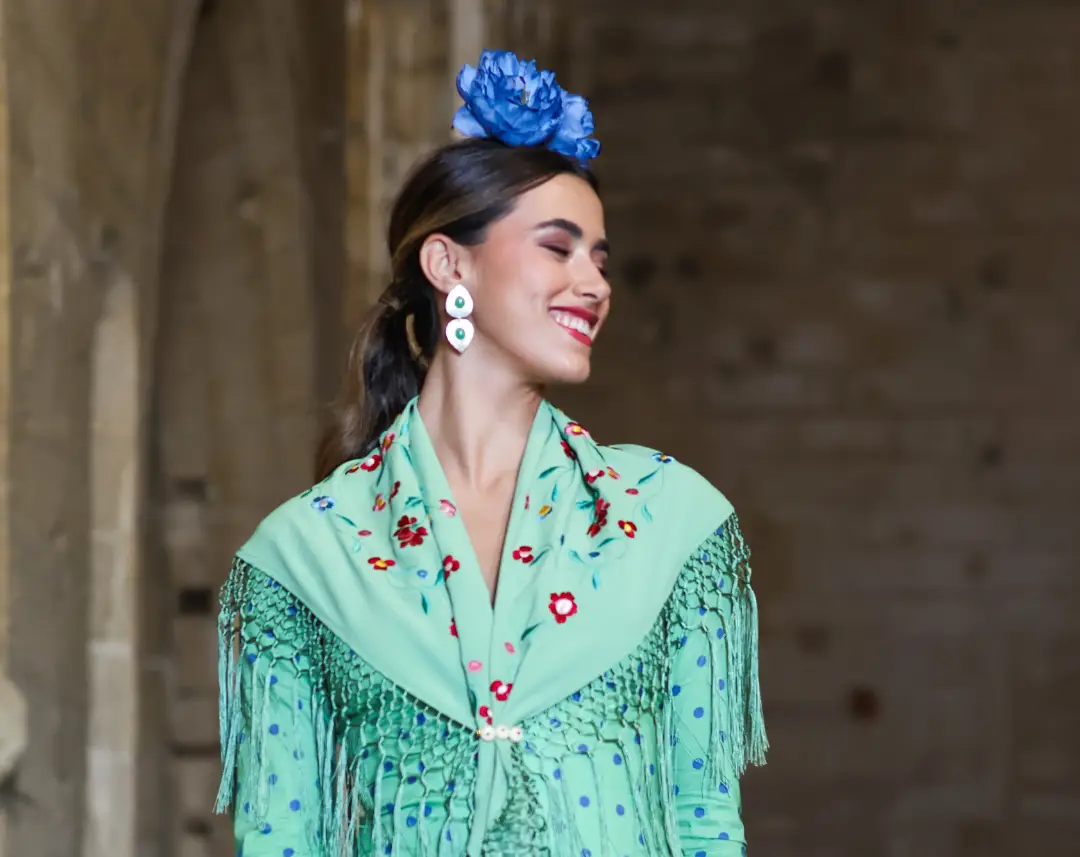 Mujer con traje de flamenca verde, flores azules en el cabello y flecos en el mantón